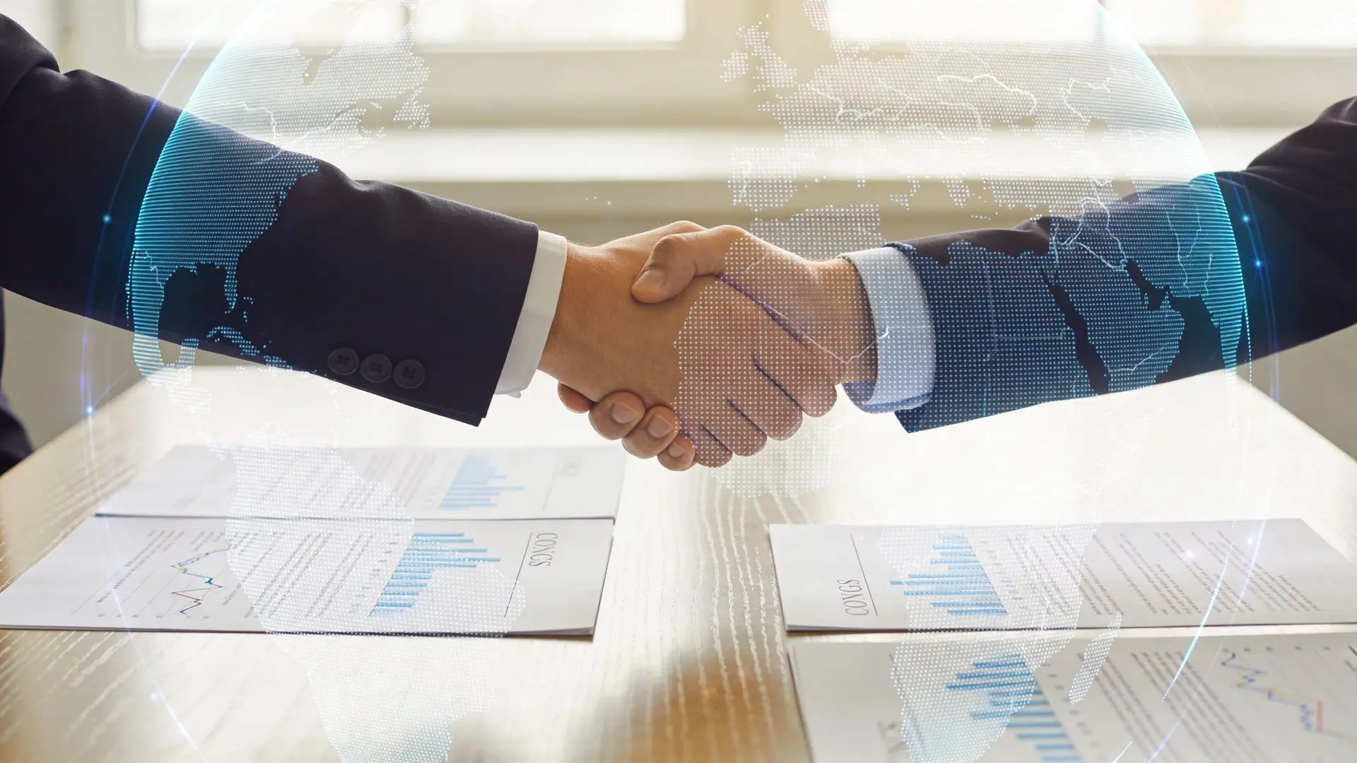 Two business people in suits shaking hands over a table with financial charts and digital globe graphics.